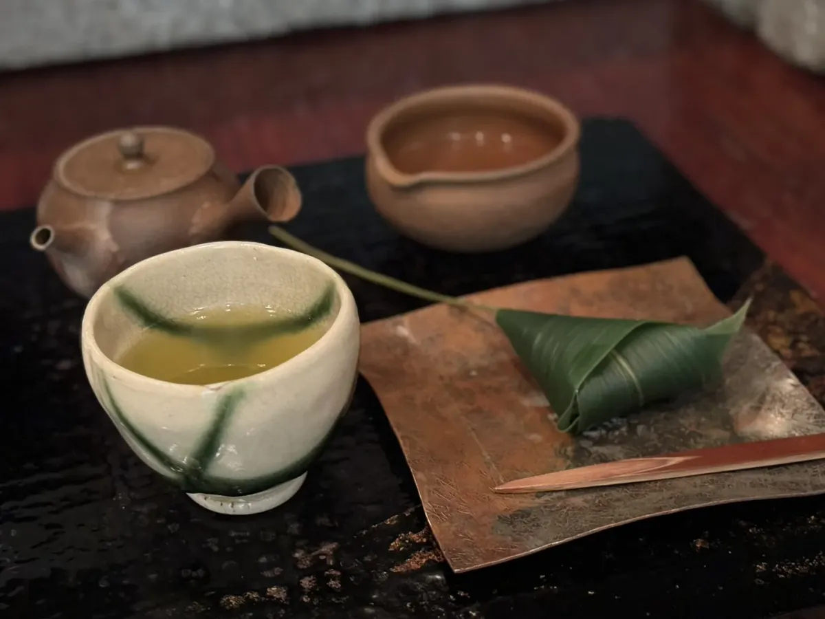 A cream and green tea bowl of pale sencha beside a small clay kyusu, a brown cooling bowl, and a sweet wrapped in a bamboo leaf on a copper plate