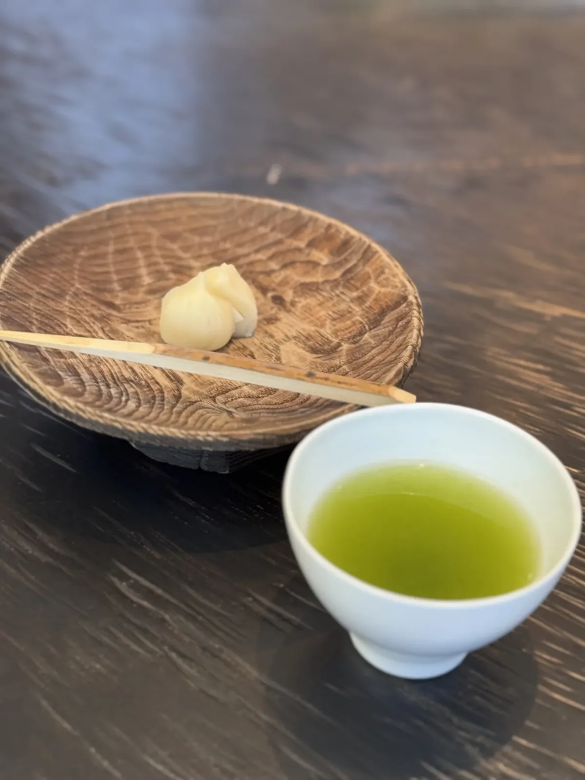 A single small white wagashi on a carved wooden plate with a bamboo pick, next to a cup of vivid green tea