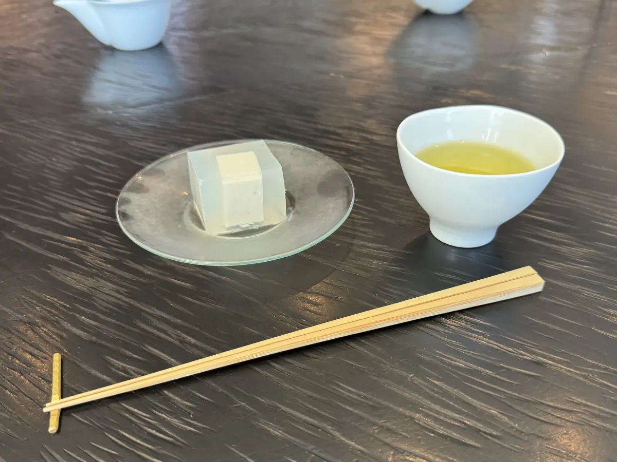 A translucent jellied sweet on a glass plate beside a cup of pale gold tea and long bamboo chopsticks