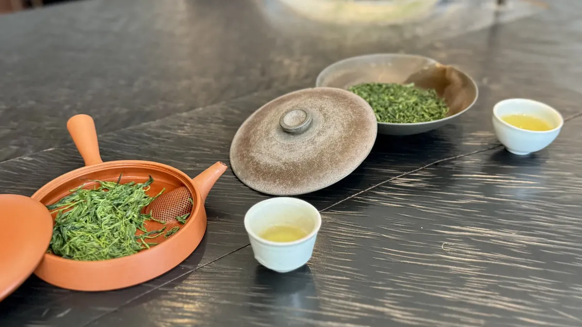A dark wood counter with a vermilion roasting vessel of fresh green tea leaves, a lidded pot, a dish of dry leaves and two small white cups of pale green tea