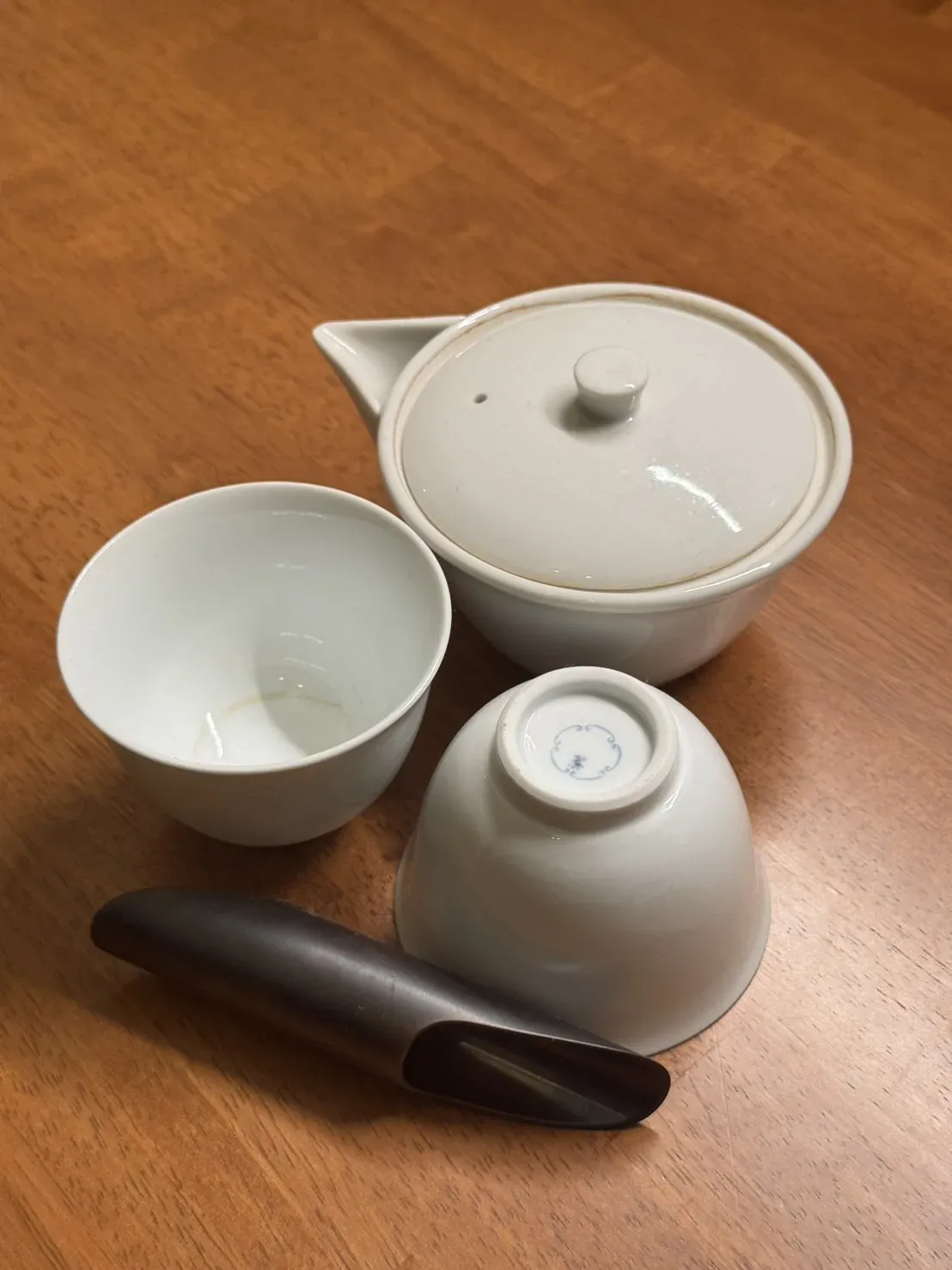 A white handleless teapot with its lid, two small white cups — one turned over to show a blue maker's mark on the foot — and a dark wooden tea scoop, on a wooden floor at home