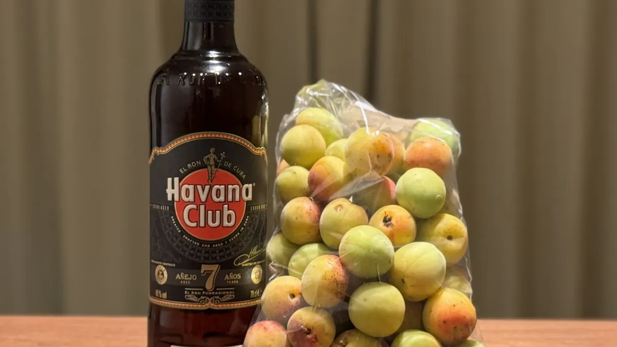 A net bag of hard green ume plums beside a bottle of aged dark rum on a wooden table