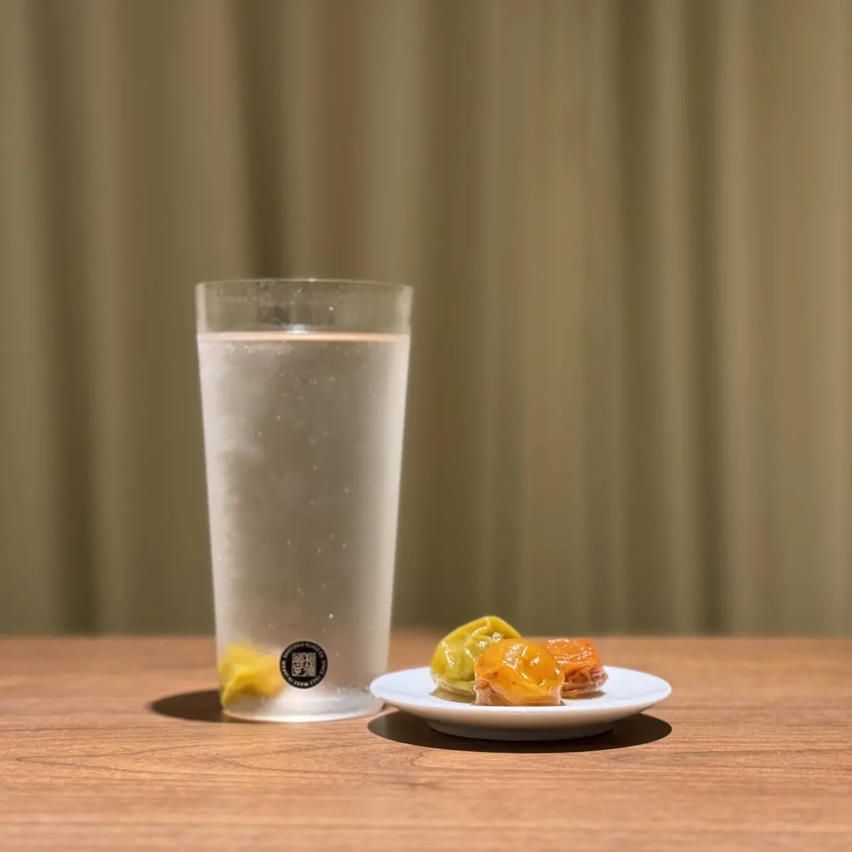A tall glass of ume syrup over ice, with a small dish of preserved plums beside it